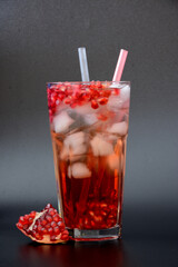 A tall faceted glass of refreshing pomegranate juice with ice and tubes on a black background, next to it is a broken ripe fruit with seeds.