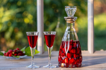 Homemade red raspberry brandy in two wine glasses and in a glass bottle on a wooden windowsill near summer garden, closeup