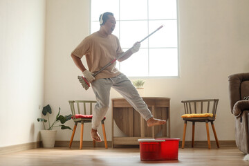 Asian man jumping doing guitarist pose while mopping floor at living room, wearing headphones and dancing with music.