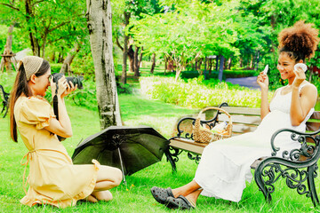 Asian female friend photographer sitting and taking shot photo, portrait healthy pregnant african american woman sitting on wooden bench in park showing gloves baby smiling looking at camera happy.