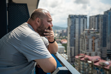 Overweight man feeling bad suffering from heat in summer standing on balcony looking at street in summer. High sweating, hyperhidrosis, obesity, heaviness from hot high humidity, excess body weight. 