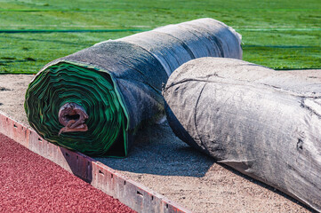 Rolls of green grass carpet and orange soft crumb rubber tracks close up. Laying artificial turf on...
