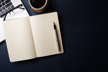 Flat lay, top view of blank notebook, pen, cup of coffee, laptop and glasses on black background.