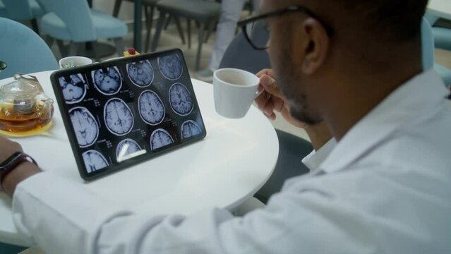 African American doctor watches image of MRI or CT scan using digital tablet in clinic cafe. Medic drinks tea, examines brain scanning results of patient. Medical staff in modern hospital cafeteria.