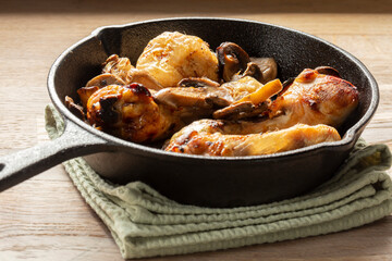 Fried chicken drumsticks and sliced mushroom n a cast iron frying pan. On a wooden chopping board in a kitchen