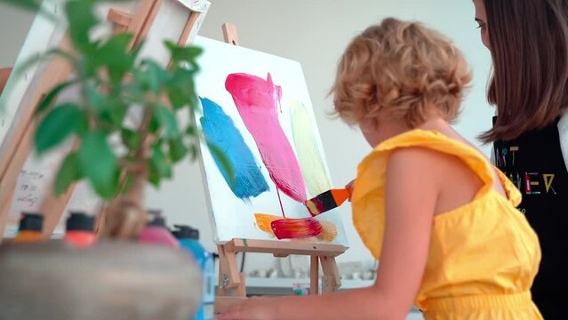 Teacher At Art School Helping Children in Art Class with painting. Woman teaches child to create and work with acrylic paint. Group of primary school kids learning how to draw and color help of their