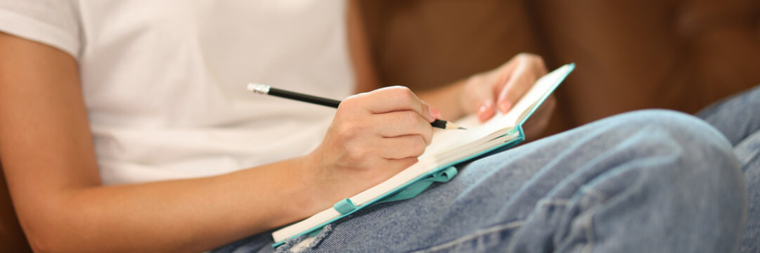 Close-up of woman taking notes in notebook while sitting on couch.