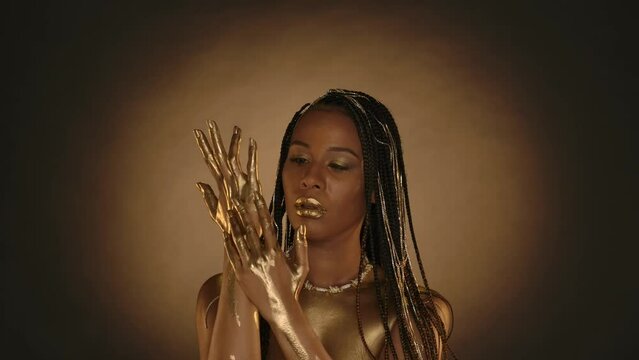 A woman with golden lips, golden skin on a brown background with circular light. A seminude African American woman applies, rubs golden paint, liquid gold into the skin of her hands.