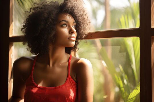 Woman wearing red tank top is seen looking out of window. This image can be used to depict curiosity, longing, or anticipation. It is suitable for various themes and concepts.