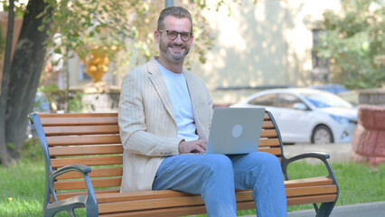 Fototapeta premium Modern Casual Man Smiling at Camera while Working on Laptop Outdoor