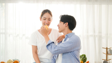 Asian young happy couple boy and girl feeding bread bakery and take care each other in the morning breakfast romantic lifestyle routine with sunrise at the window curtain in a kitchen apartment