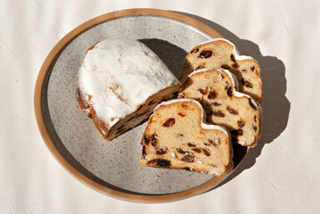 Christstollen, traditional holiday German bakery. Fruit bread or cake with raisins and sugar powder on plate, on beige table background in sun light.
