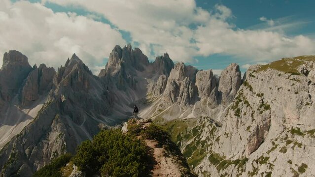 Parralax drone shot with lone tourist watching panoramic view with tall steep rocky mountains, partly clouded sky in the background, hiking in the Alps, majestic landscape,  cinematic color grade