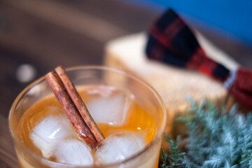 A glass of whiskey with ice and a cinnamon stick. A juniper branch and a checkered bow in the...