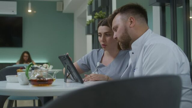 Diverse doctors sit in modern clinic cafe. Professional medics talk, watch medical tests results using digital tablet, discuss diagnosis. Medical staff work in hospital or medical center cafeteria.