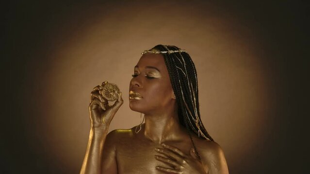 A woman in Cleopatra style on brown background with circular light. A woman with skin in golden paint holds a half of a lemon. Lemon juice flows down the woman's hand. Slow motion.