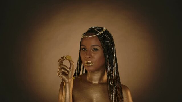 A woman in Cleopatra style on brown background with circular light. A woman with golden skin, with jewelry on her head, eats a golden candy. Fashion art design. Slow motion