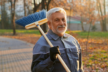 Smiling maintenance male worker looking at camera, while keeping broom on shoulder. Portrait of bearded man in age, wearing uniform standing with broom, against natural background. Workplace concept. 