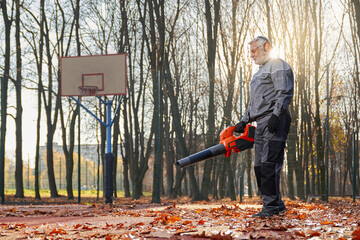 Smiling janitor with grey hair cleaning autumn city park with leaf blower. Side view of senior man...