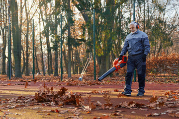 Senior man in overalls, headphones and gloves cleaning fenced city park area from dry fall leaves....