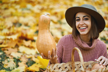 Close up autumn portrait of happy smiling lady in hat holding big rape pumpkins outdoors, fall season and colorful foliage around on backdrop