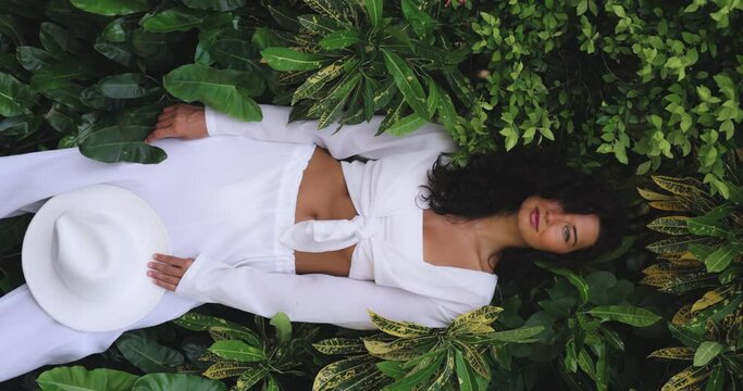 A beautiful tanned fashionable girl with natural make-up and hairstyle stands in white hat and white stylish clothes among exotic tropical plants. High quality 4k slow motion vertical video