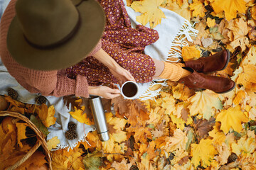 Top view of fashionable faceless woman in hat enjoying coffee in autumn park, surrounded by colorful leaves