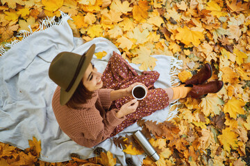 Top view of joyful stylish woman in hat holding cup of coffee, enjoying picnic time at park covered of foliage. Focus on cup