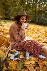 Elegance young lady in hat enjoying of coffee at autumn park, while sitting in blanket surrounded orange leaves