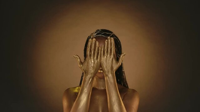 A woman with creative golden make up and golden skin on a brown background with circular light. A woman covers her face with her palms, runs her hands down her neck and to her shoulders. Slow motion.