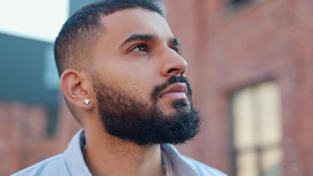 Close up view of serious handsome bearded man with little earring dressed in blue shirt looking at sky and dreaming. Young pensive man of arabian nationality thinking of own purposes in life.