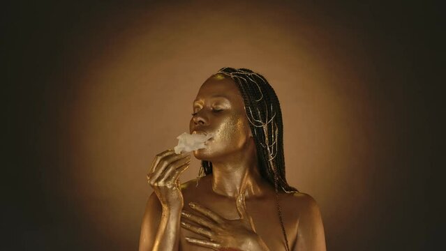 A seminude woman with golden skin touches herself with a bud of a white gladiolus flower. Woman with golden metallic skin in studio on brown background with circular light. Fashion, art. Slow motion.