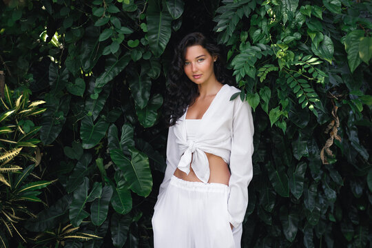 Natural beauty portrait of fashionable girl with natural make-up and hairstyle stands in white stylish clothes among exotic tropical plants. High quality 4k slow motion footage 