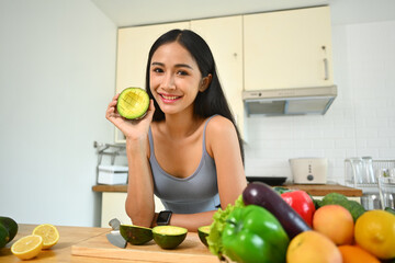 Happy sportswoman holding avocado, preparing ingredients for making healthy vegetable detox smoothie in kitchen
