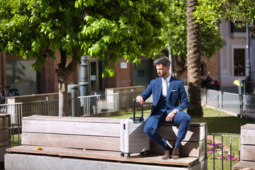 Obraz premium Young and attractive businessman, with beard and suit, sitting on a bench next to his suitcase waiting for a cab. Concept beauty, fashion, success, achiever, trend.