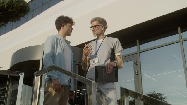 Mature doctor with digital tablet in hands stands with adult patient at clinic entrance and discuss his treatment. Health care specialist consults client or makes diagnosis. Physician works outside.