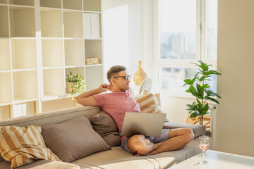 Young attractive Asian man relaxing, stretching on sofa in apartment with laptop on lap, remote work and freelancer concept