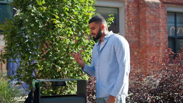 Casually dressed bearded man of indian ethnicity strolling on fresh air, breaking cigarette and throwing it in street trash can. Conscious adult giving up smoking and encouraging to healthy lifestyle.
