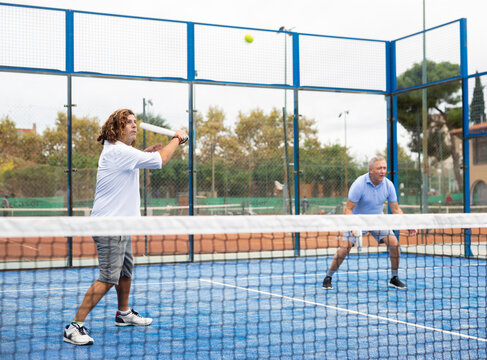 Portrait of adult man enjoying padel racket sport, playing doubles game with elderly male friend on court outdoor