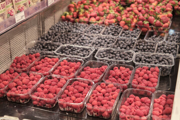 plastic trays filled with glistening raspberries and blueberries. rows of berries symbolize the paradox of modern life: need for convenience over sustainability, berry in single-use plastic.