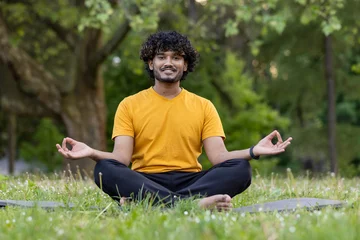 Fotobehang Indian young man doing yoga in the park. Sitting on a sports mat in the lotus position and meditating relaxed. He looks at the camera with a smile © Tetiana