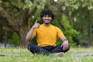Gordijnen A young Indian man does sports and yoga in the park. Sitting on the mat in the lotus position, smiling at the camera and pointing super © Tetiana