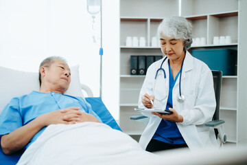 Asian doctor in white suit take notes while discussing and Asian elderly, man patient who lying on bed with receiving saline solution in hospital or clinic.