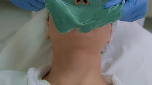 Cosmetologist removes alginate mask from female patient face, close-up. Facial skin care, anti-aging procedures in beauty salon.