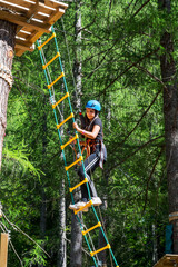 Happy young woman climbing ropes ladder in forest adventure park