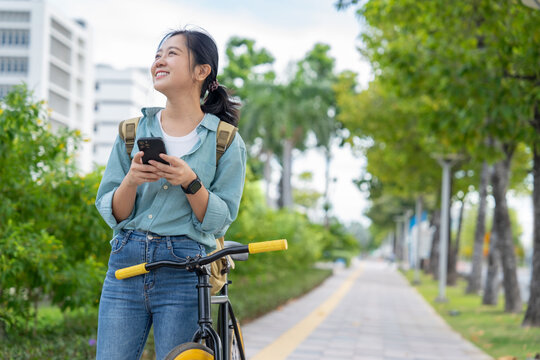 Young beautiful Asian woman is using a smartphone while riding a bicycle through a city park. Lifestyle and environmentally friendly concept. - Powered by Adobe