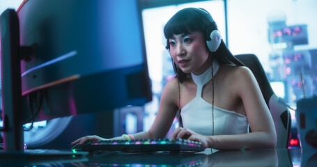 Enthusiastic Asian Girl in Headphones Talking with Friends Online on a Computer. Stylish Streamer or Video Gamer Playing Online Simulator and Chatting with Internet Followers in a Futuristic Studio