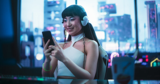 Beautiful Smiling Asian Female Using Smartphone while Sitting Behind a Desk in a Futuristic Cyberpunk Office Space. Young Japanese Female is Connecting with Internet Friends, Browsing Online Shops