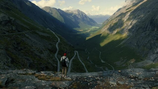 Drone flies over a young man tourist standing on the edge of a fjord towards an epic panorama of a Trolls path road