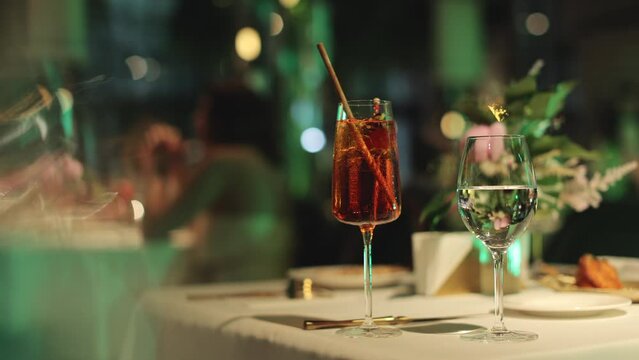 Close up of round table with white tablecloth being served with tableware and decorated with flowers for special event in restaurant. Two glasses with drinks placed on edge.